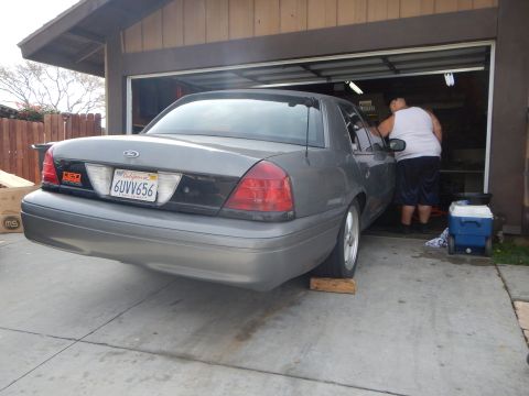 2003 Ford Crown Victoria - gray Ford sedan is backed into a garage with its rear wheels on wooden ramps while a person works at the open driver door.