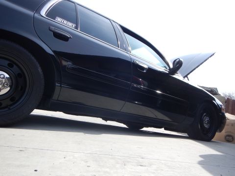 2003 Ford Crown Victoria - Dramatic low-angle side shot of a black Ford Crown Victoria sedan with black steel wheels and an open hood on a concrete surface.