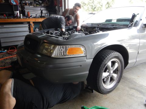 2003 Ford Crown Victoria - Two men lean over the engine bay of a gray Ford Crown Victoria with its hood up while another person works at the front wheel.