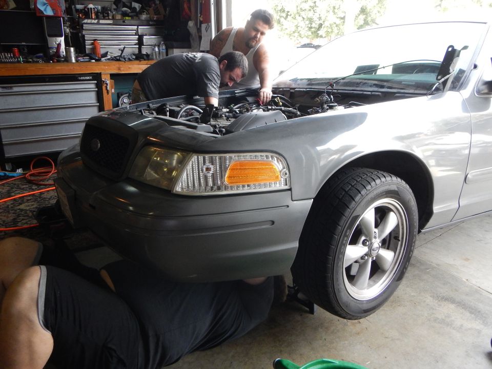 Two men working under the hood of a gray Ford Crown Victoria in a garage