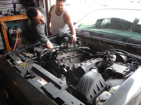 2003 Ford Crown Victoria - Two men lean over the exposed V8 engine bay of a Ford sedan in a garage, with the air intake and battery visible.