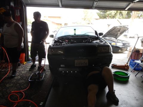 2003 Ford Crown Victoria - Men work on a dark Ford sedan with its hood up in a backlit garage, one lying under the front bumper and others standing nearby.
