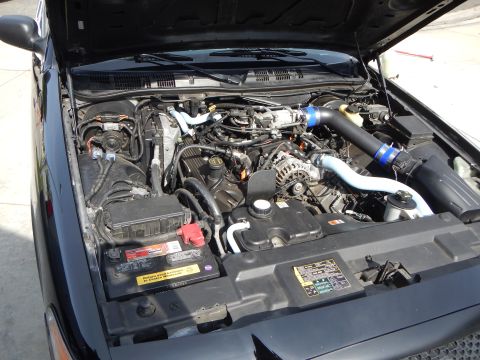 2003 Ford Crown Victoria - Close-up of a Ford sedan's engine bay showing the V8, alternator, battery, and a modified intake with blue and black piping.