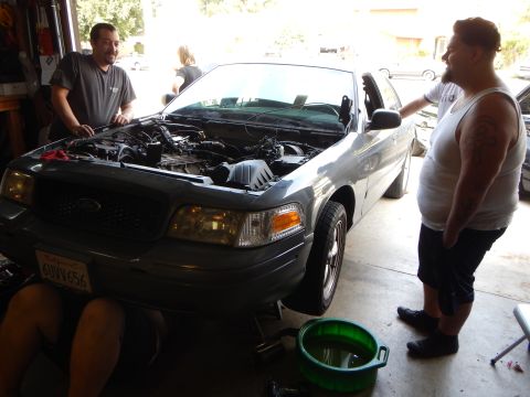 2003 Ford Crown Victoria - Two men converse beside a silver Ford Crown Victoria sedan with its hood up and an oil drain pan on the garage floor.