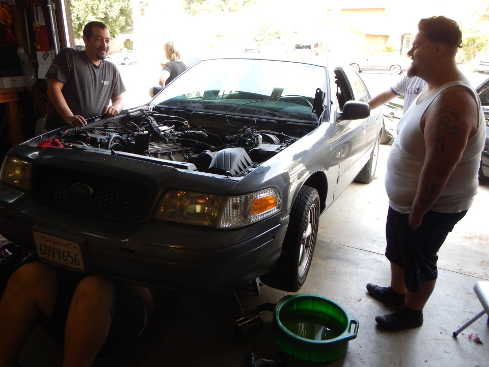Two men talking beside a silver Ford sedan with hood open in a garage, drain pan on floor