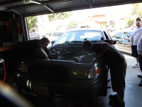 2003 Ford Crown Victoria - Two men work under the hood of a dark Ford sedan in a garage while a bystander watches at right.