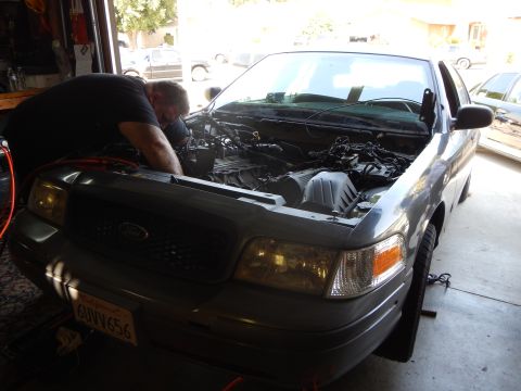 2003 Ford Crown Victoria - man bends over the open engine bay of a silver Ford Crown Victoria sedan parked in a garage doorway.