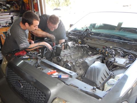 2003 Ford Crown Victoria - Two men lean into the engine bay of a vehicle in a garage, working together on the engine with tools laid out.