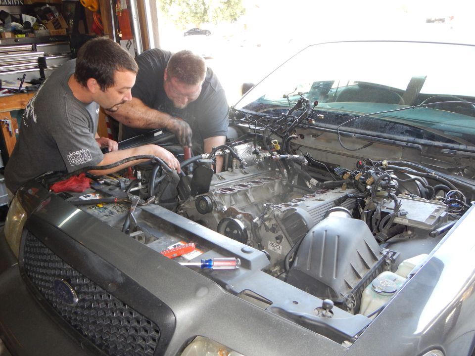 Two men leaning into the engine bay of a vehicle in a garage working on the engine