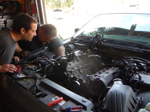2003 Ford Crown Victoria - Two men work over the open engine bay of a dark vehicle in a garage with tools spread across the front.