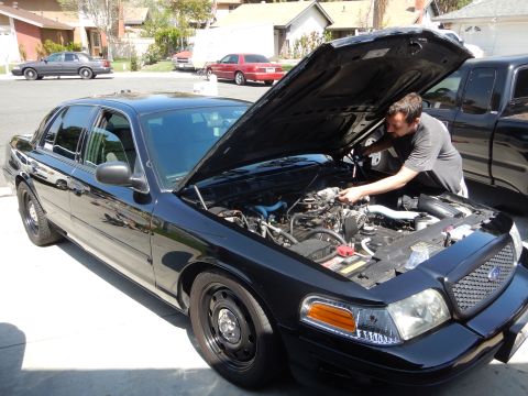 2003 Ford Crown Victoria - man works under the open hood of a black Ford Crown Victoria sedan parked in a residential driveway.