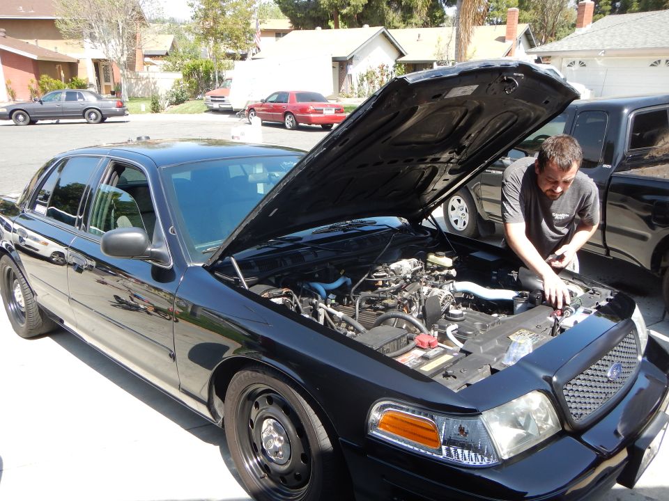 Black Ford sedan with hood up while a man reaches into the engine bay in a driveway