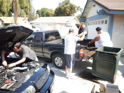 2003 Ford Crown Victoria - driveway scene with a black sedan undergoing engine work in the foreground while men unload mulch from a black pickup.
