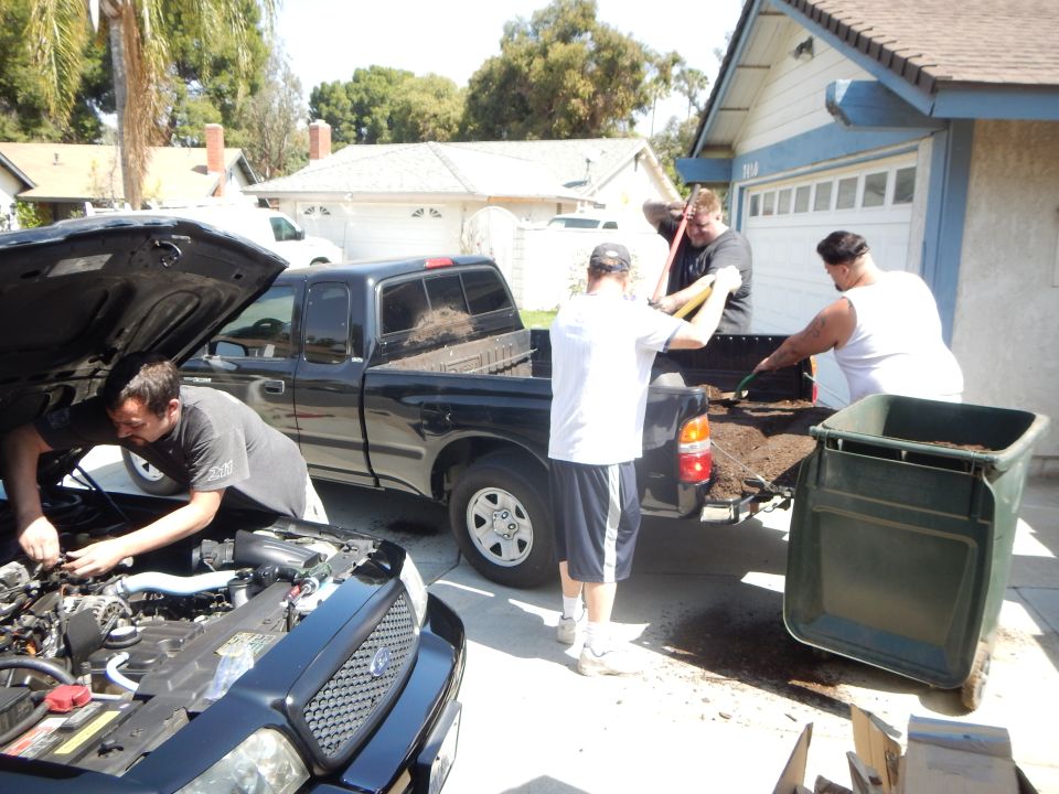 Driveway scene with a black sedan having engine work and men unloading a black pickup of soil