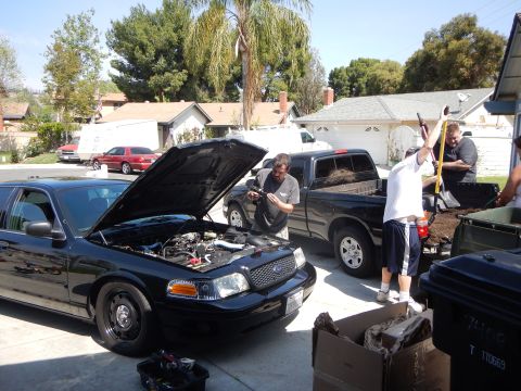 2003 Ford Crown Victoria - black Ford Crown Victoria sedan with open hood sits beside a black pickup as men unload mulch in a residential driveway.