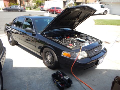 2003 Ford Crown Victoria - black Ford Crown Victoria sedan sits with its hood up and a red jumper lead running to the engine in a driveway.