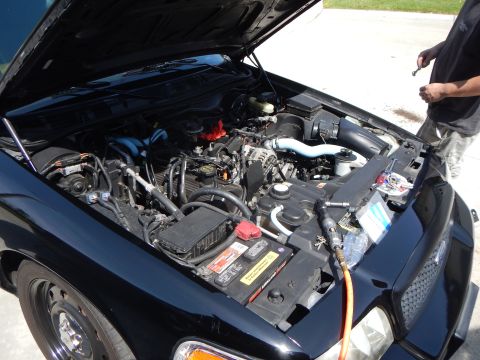 2003 Ford Crown Victoria - Close-up of a black Ford sedan's engine bay showing the battery and a yellow hose, with a man standing at right.