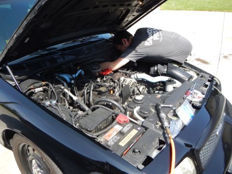 2003 Ford Crown Victoria - man bends into the engine bay of a black Ford sedan, working on the engine with a yellow hose connected, in a driveway.