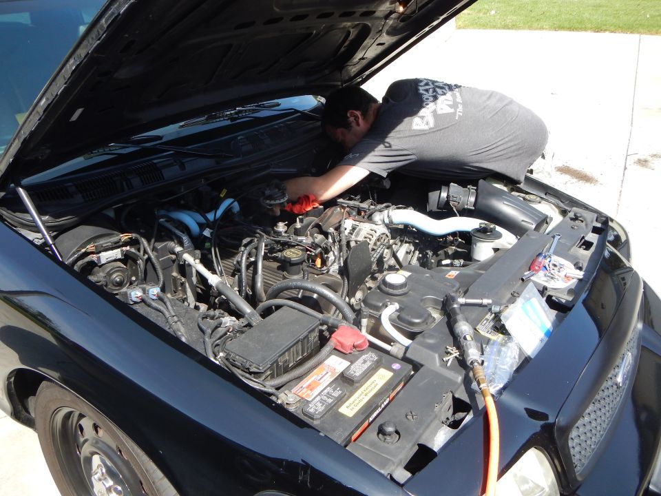 Man bent into the engine bay of a black Ford sedan working on the engine in a driveway