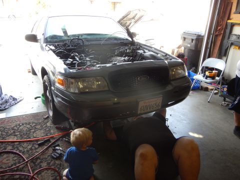 2003 Ford Crown Victoria - silver Ford Crown Victoria sedan sits with hood up in a garage while a man works underneath and a toddler stands by.