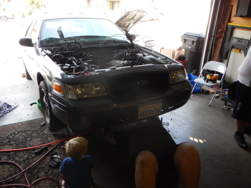 Silver Ford sedan with open hood in a garage, person under it and a toddler nearby