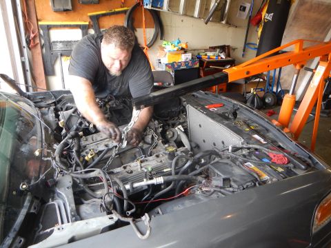 2003 Ford Crown Victoria - man works on a disassembled engine in a vehicle's bay using a chain, with an orange engine hoist in the garage behind.