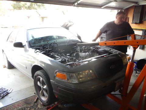 2003 Ford Crown Victoria - man leans into the open engine bay of a gray Ford sedan on jack stands inside a garage, beside an orange engine hoist.