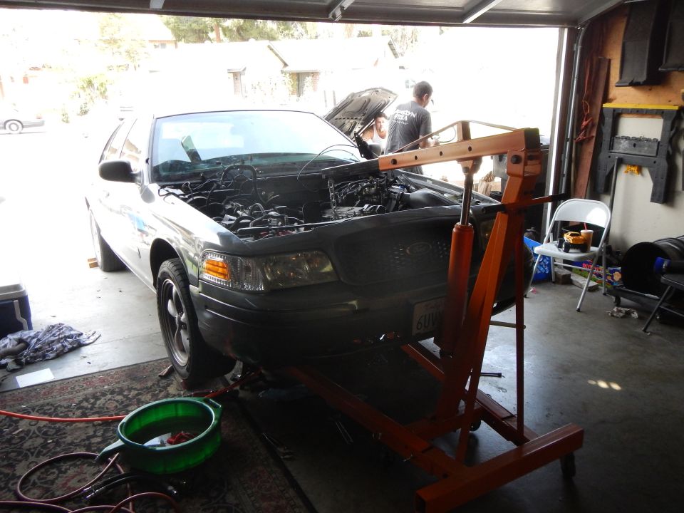 Gray Ford sedan with hood up and orange engine hoist positioned at the front in a garage