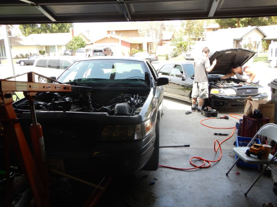 Garage with a gray Ford sedan undergoing engine work and a second vehicle outside the door