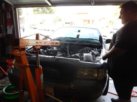 2003 Ford Crown Victoria - man stands at the fender of a gray Ford sedan whose engine bay is opened under an orange engine hoist boom.