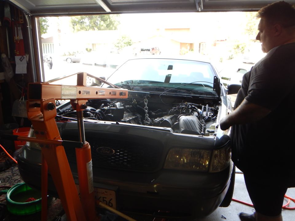 Man stands beside a gray Ford sedan with its engine partly removed under an engine hoist
