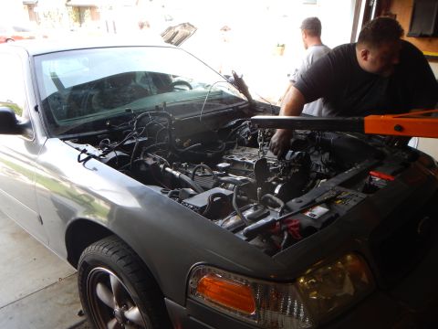 2003 Ford Crown Victoria - Two men lean over the open engine bay of a gray Ford sedan, with exposed wiring and an orange hoist arm above.
