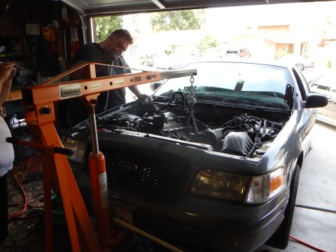 2003 Ford Crown Victoria - man stands behind an orange engine hoist at the front of a gray Ford sedan, its grille bearing the Ford oval emblem.