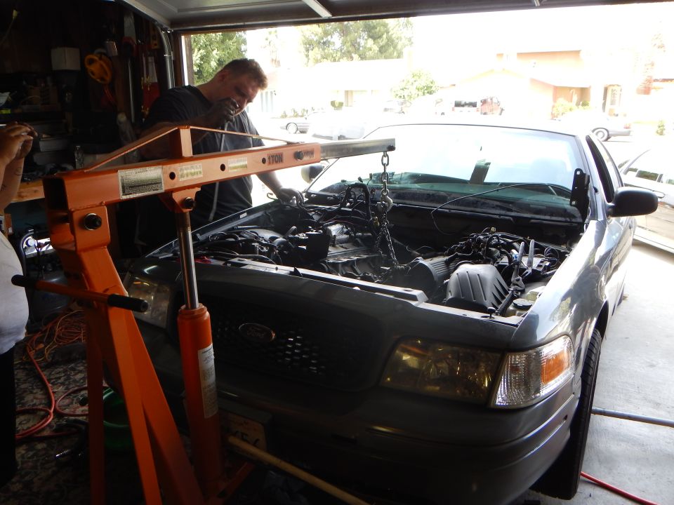 Man behind orange engine hoist working at the front of a gray Ford sedan, Ford grille emblem visible