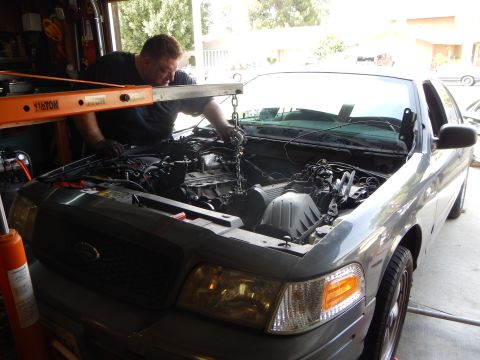 2003 Ford Crown Victoria - man reaches into the engine bay of a gray Ford sedan beneath an orange hoist labeled 1.5 ton in a garage.