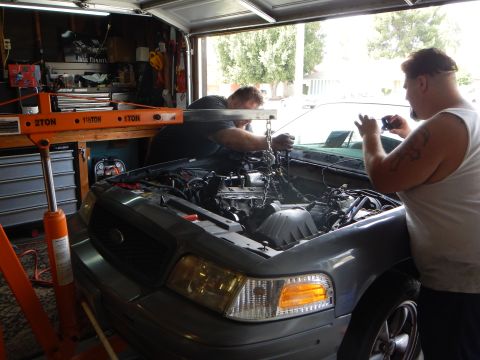 2003 Ford Crown Victoria - Two men work on a gray Ford sedan's engine bay while a tattooed man stands beside it holding a phone, near an orange hoist.