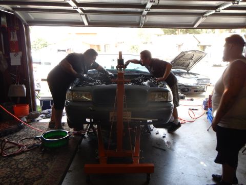 2003 Ford Crown Victoria - Three people gather around the front of a gray Ford sedan during engine work, with an orange hoist and green pan nearby.