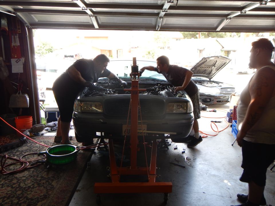 Three people gather around the front of a gray Ford sedan during engine work in a garage