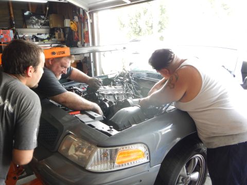 2003 Ford Crown Victoria - Three men lean over the front engine bay of a gray Ford sedan, one tattooed in a white tank top, working among the wiring.