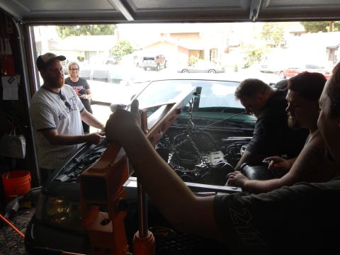 2003 Ford Crown Victoria - Several people crowd around the open engine bay of a gray Ford sedan in a garage, with an orange hoist arm in the foreground.