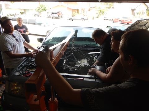 2003 Ford Crown Victoria - group works on a gray Ford sedan with its engine partly removed and lifting chains hanging into the bay.