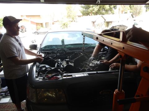 2003 Ford Crown Victoria - man in a cap watches others hoist the engine out of a gray Ford sedan using chains and an orange engine hoist.