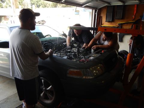 2003 Ford Crown Victoria - Three people work on the open engine bay of a gray Ford sedan parked beside a silver minivan inside a garage.