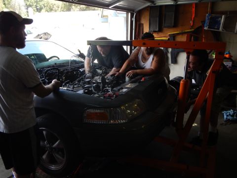 2003 Ford Crown Victoria - Three people lean into the engine bay of a gray Ford sedan, its grille showing the Ford oval, beneath an orange hoist.