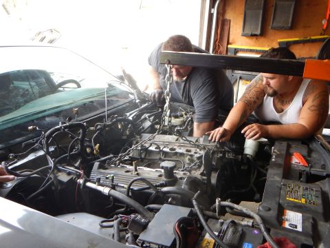 2003 Ford Crown Victoria - Two men work over an exposed V-style engine in the bay of a gray Ford sedan, with wiring and a Ford service label visible.