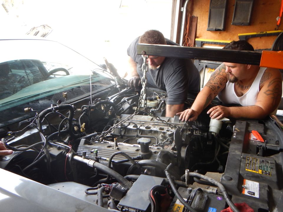 Two men work over an exposed V8 engine in the bay of a gray Ford sedan