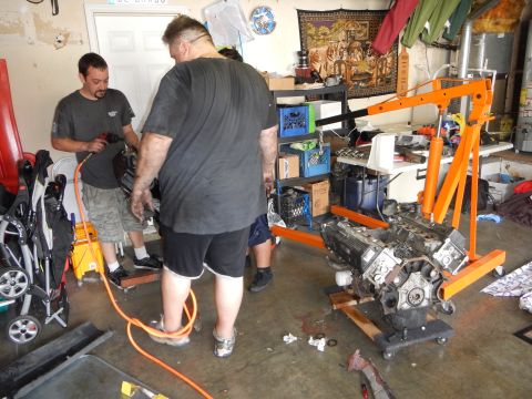 2003 Ford Crown Victoria - Men work in a cluttered garage where a V8 long block sits on an orange engine stand among scattered tools.