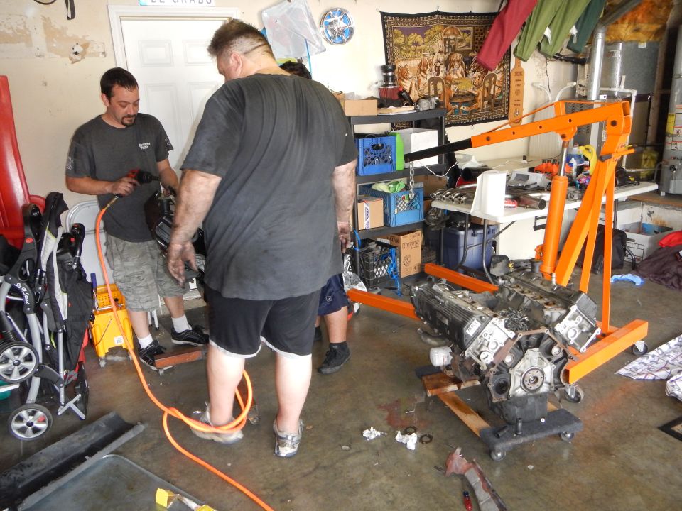 Men in a garage with a V8 long block on an orange engine stand and tools on the floor