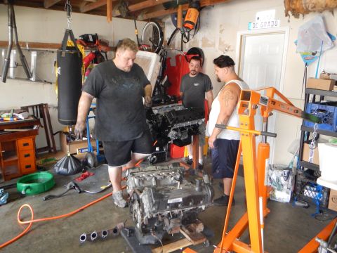 2003 Ford Crown Victoria - Three men stand around a V8 engine mounted on an orange stand in a garage with a hanging punching bag.
