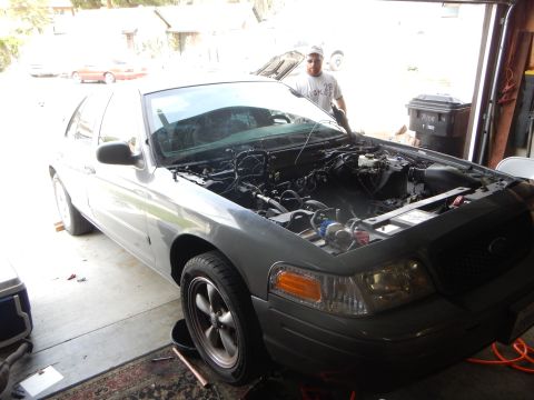 2003 Ford Crown Victoria - gray Ford sedan with its hood up and empty engine bay sits in an open garage doorway as a man stands behind it.